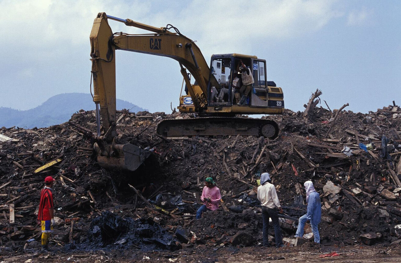 Una excavadora amarilla despeja una gran pila de escombros mientras cuatro personas ayudan, como parte de las labores de recuperación tras el tsunami en Indonesia, apoyadas por el PNUD.