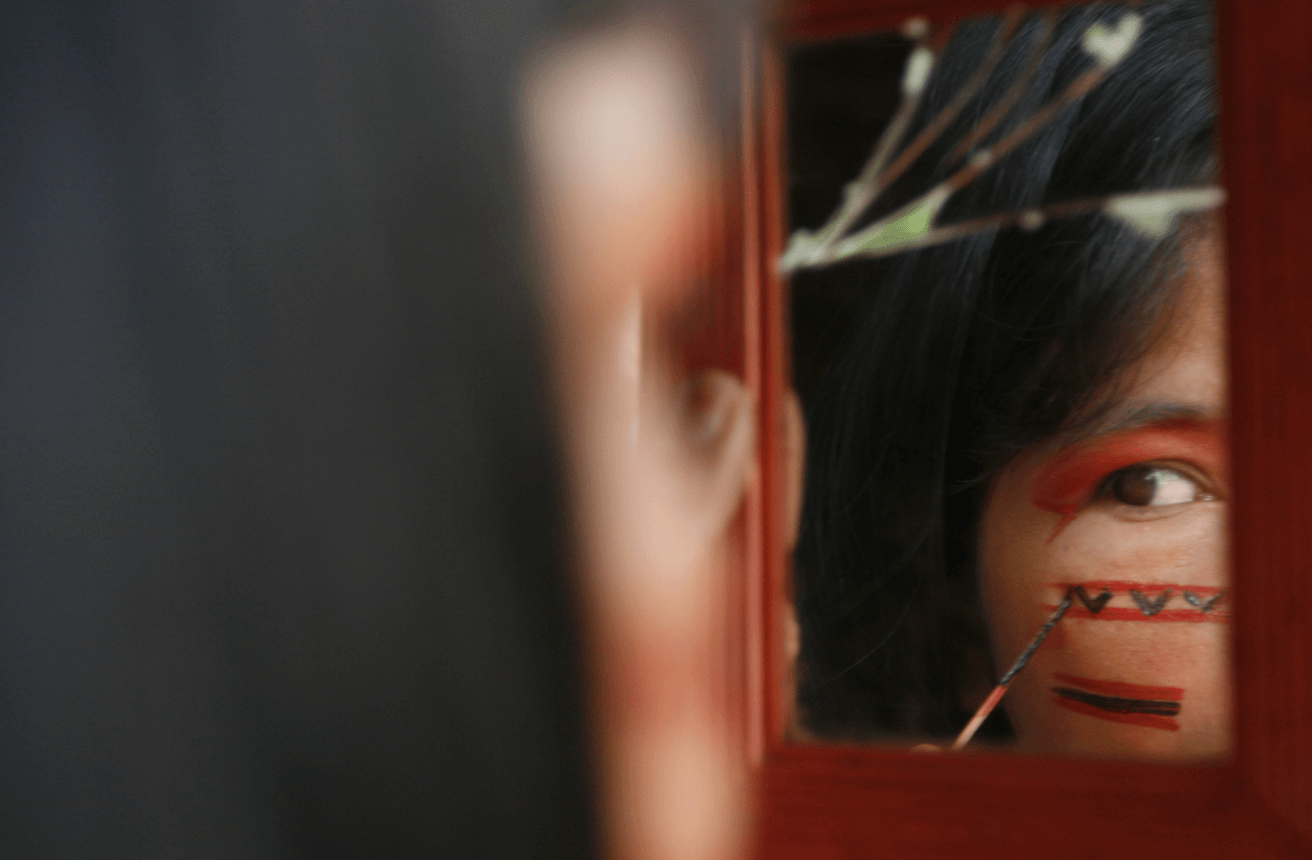 Una mujer Apiwtxa en Brasil pintandose el rostro con patrones tradicionales rojos y negros, mientras se mira en un espejo.
